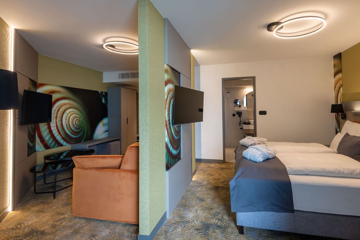 Ein modernes Hotelzimmer mit einer Couch, einem Bett, einem Fernseher und einem durchgehenden Türspalte sichtbaren Badezimmer.