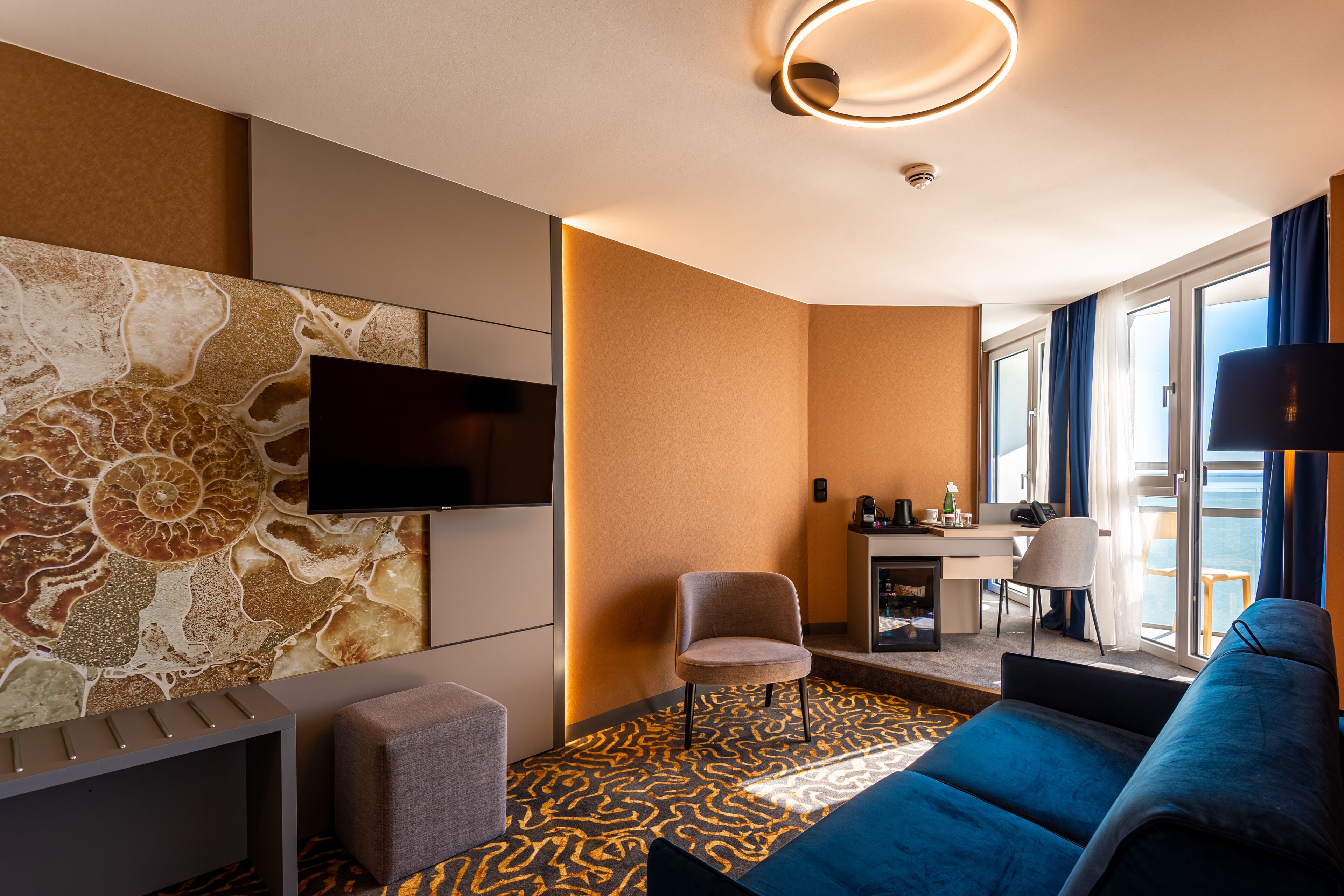 A modern hotel room with a large TV, a sofa, a desk with a chair, and a view of the sea through the windows.