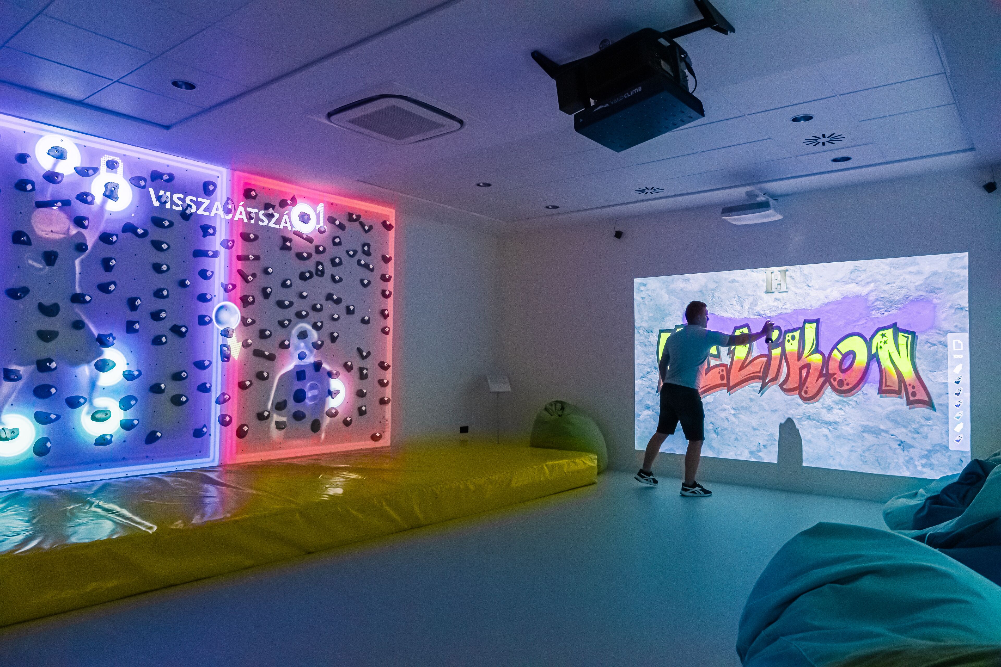 Une pièce équipée d’un mur d’escalade, d’un écran de projection et d’un espace rembourré jaune.
