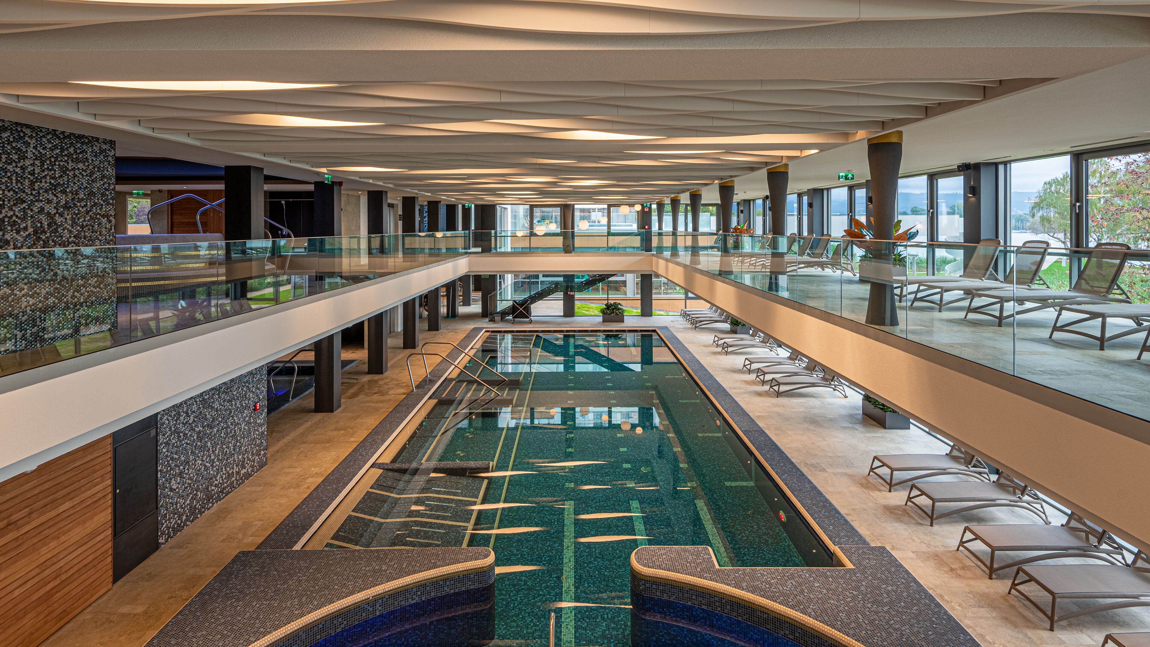 A modern indoor swimming pool with a glass barrier, lounge chairs, and a view of the outdoors.