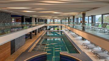 A modern indoor swimming pool with a glass barrier, lounge chairs, and a view of the outdoors.