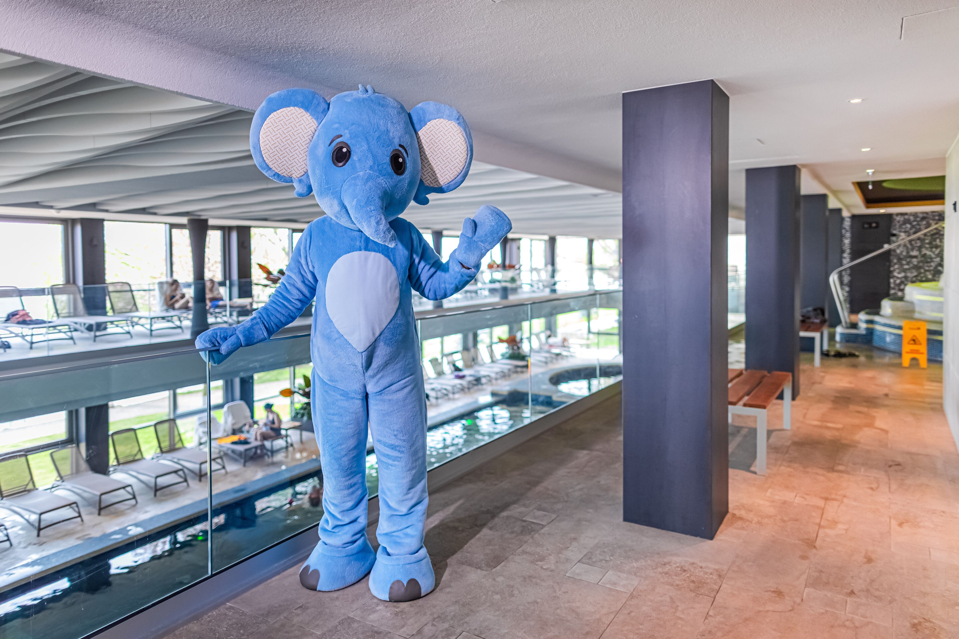 A large blue elephant mascot standing in a modern indoor space with a glass railing overlooking a pool area.