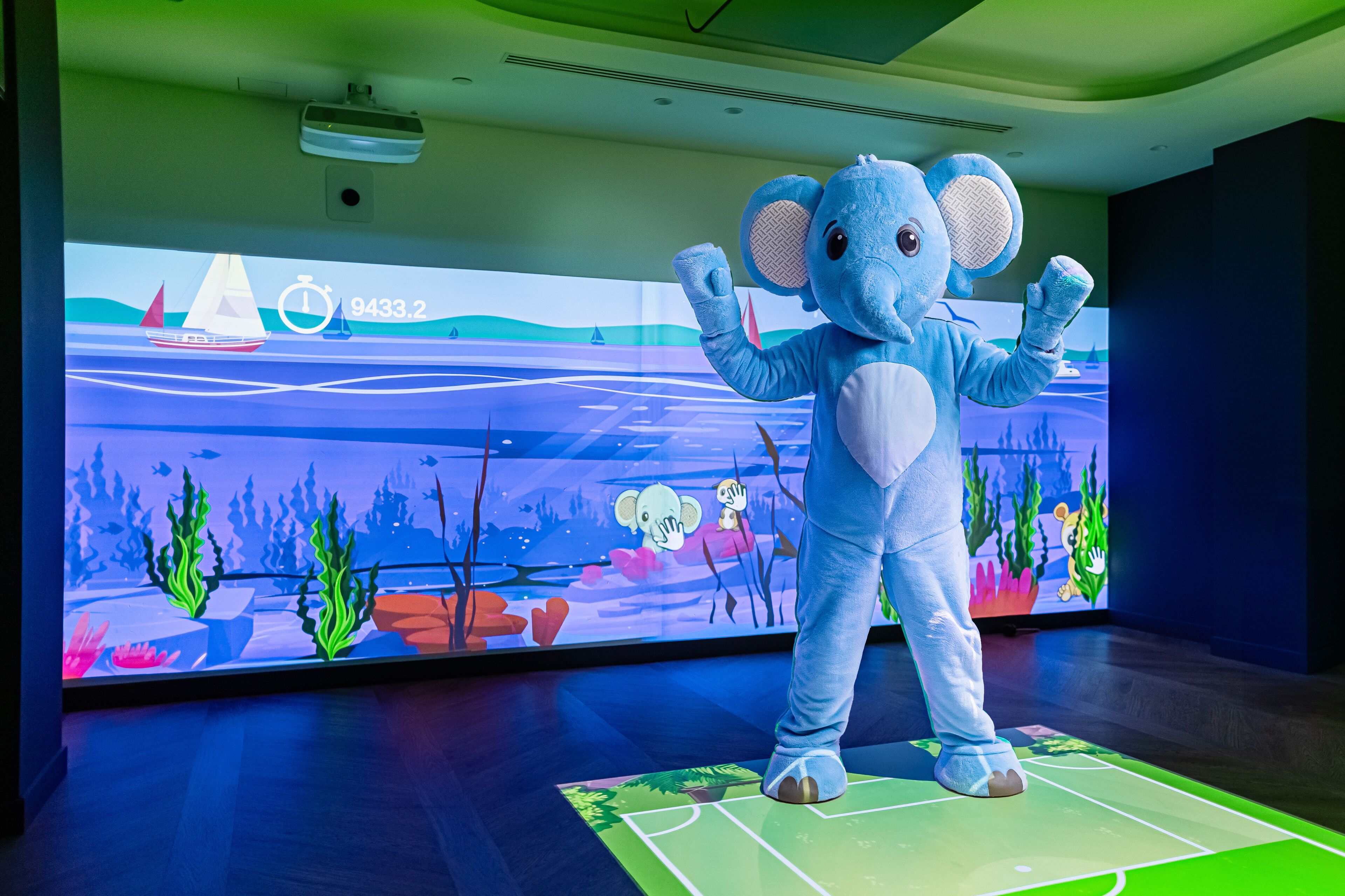 A mascot in a blue elephant costume standing on a mat in front of a large digital screen displaying an underwater scene with a sailboat.