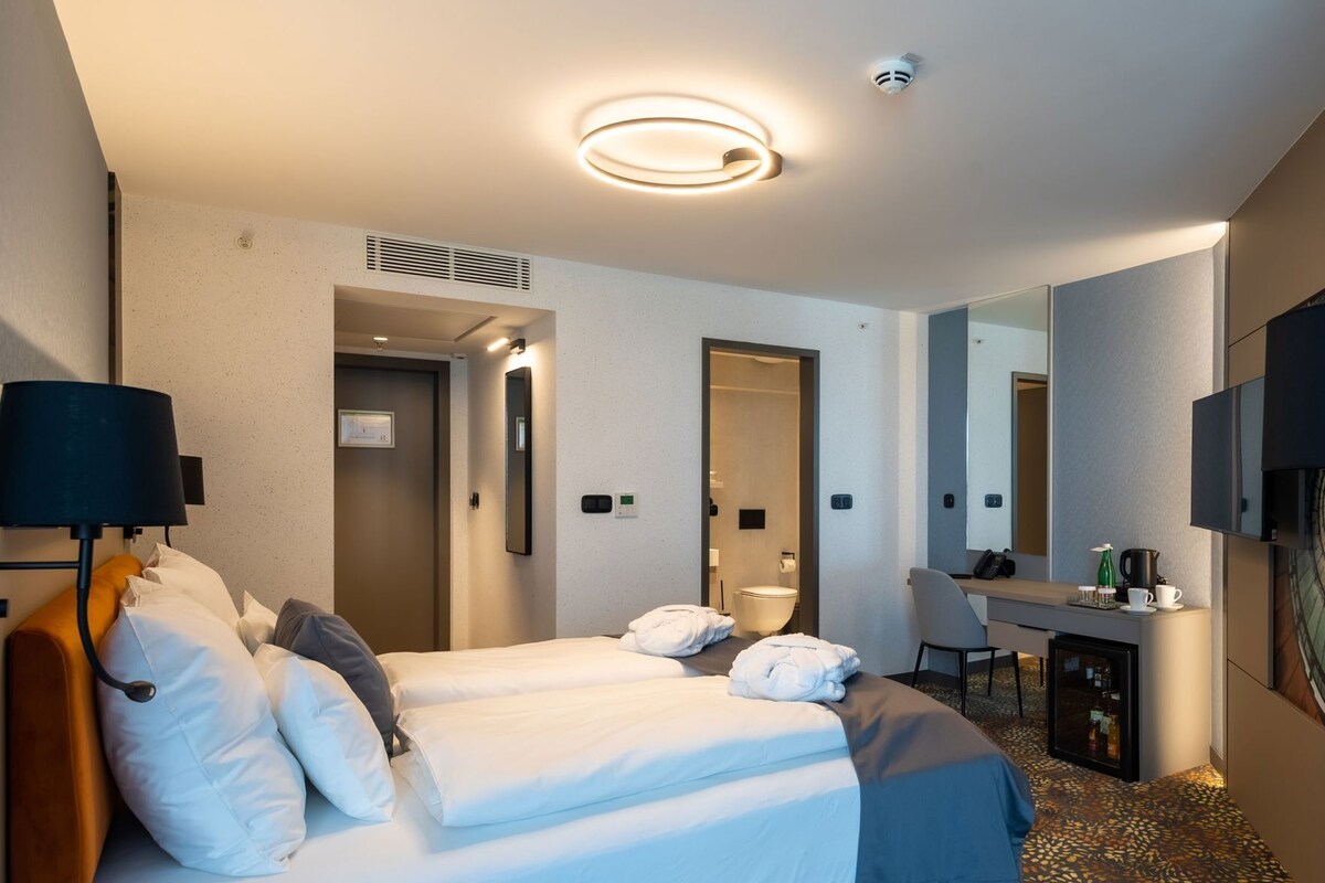A hotel room with two beds, a desk, a chair, a TV, and a minibar.