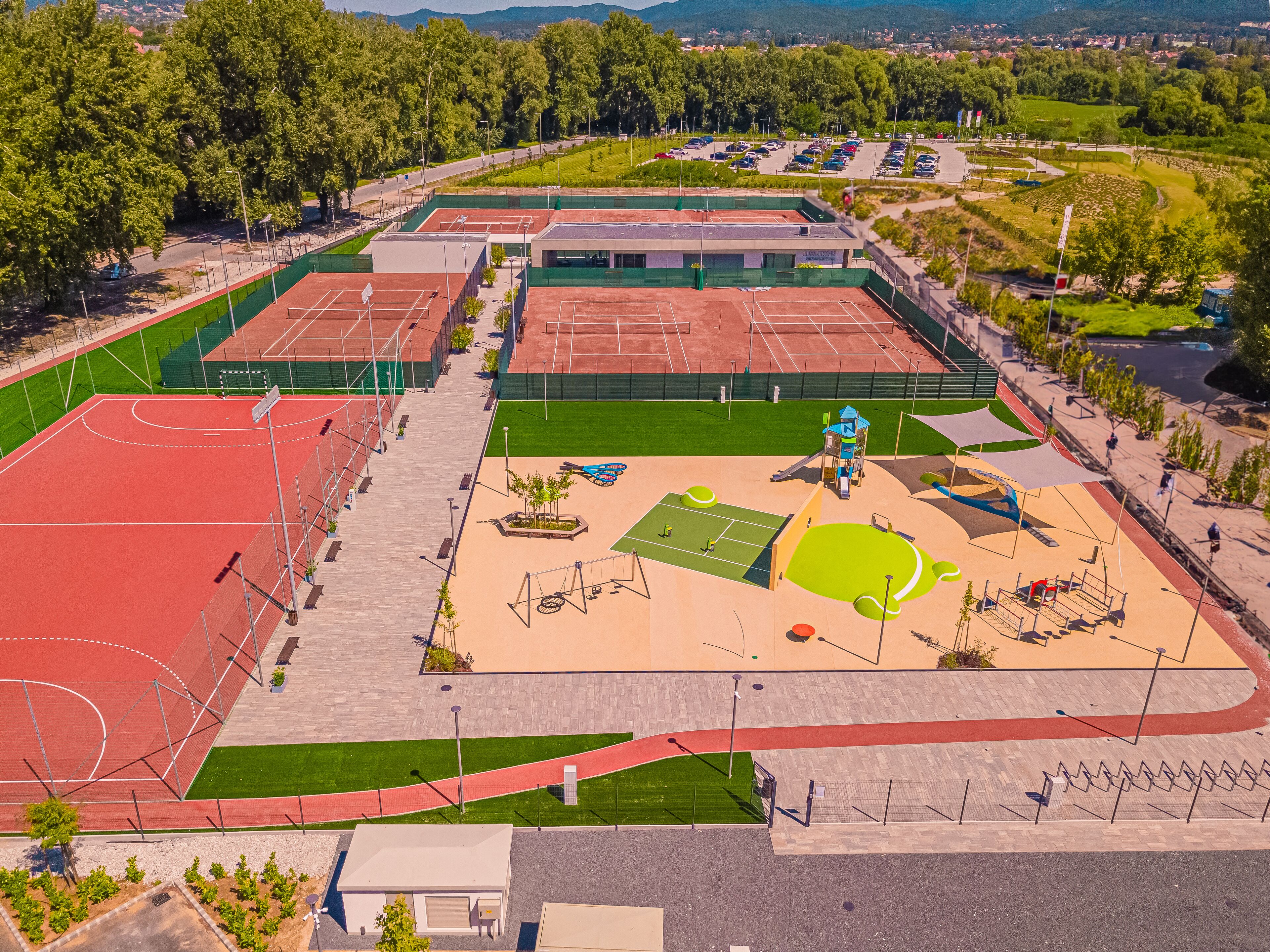 An aerial view of a sports complex with multiple tennis courts, a playground, and a parking area.