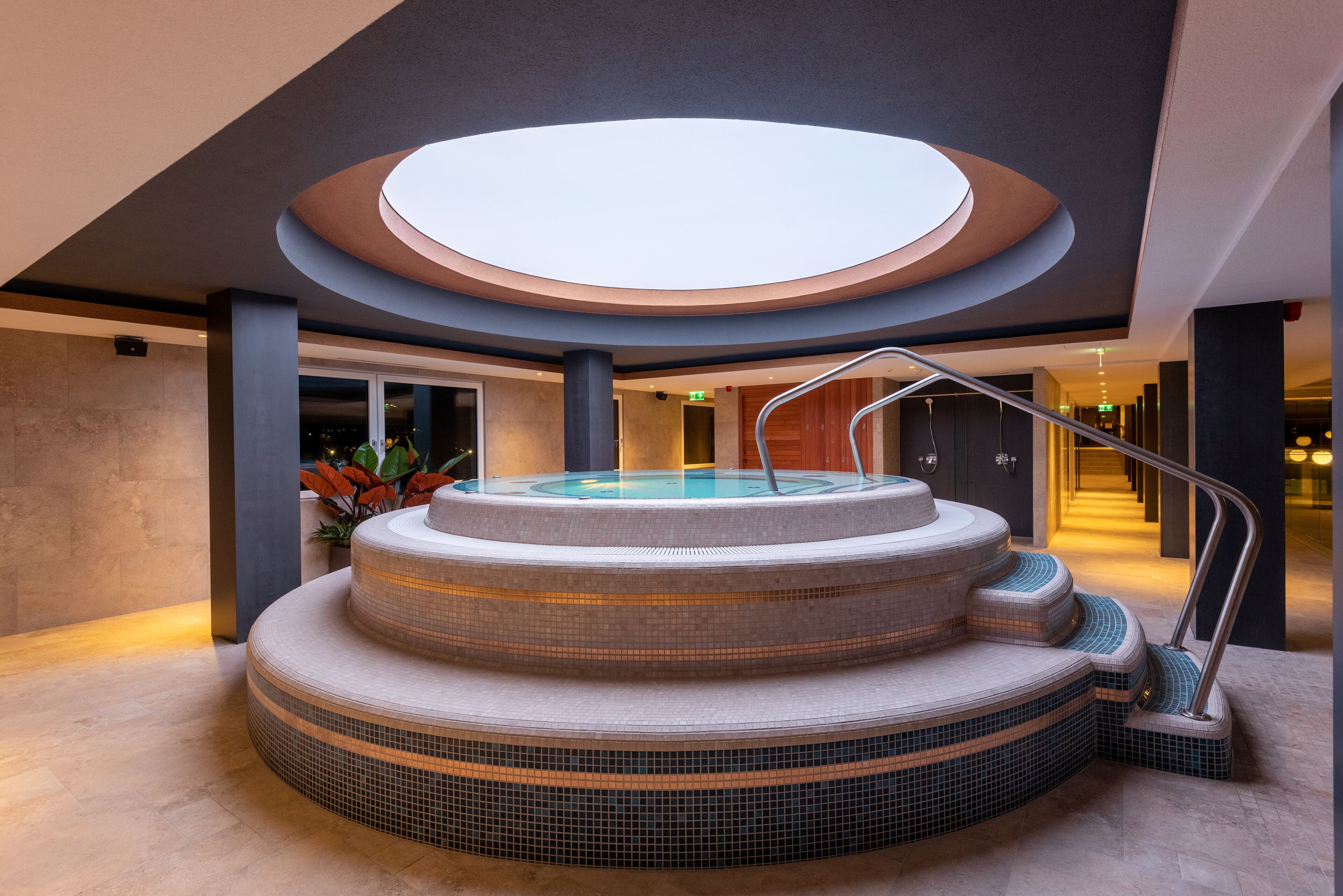Un jacuzzi de varios niveles con un tragaluz circular encima, rodeado de un suelo de baldosas y una escalera que sube.