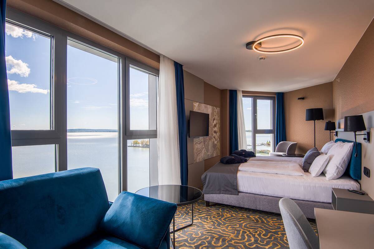 Ein Hotelzimmer mit großem Fenster, einem Bett, einem Schreibtisch, einem Stuhl und einem Fernseher.