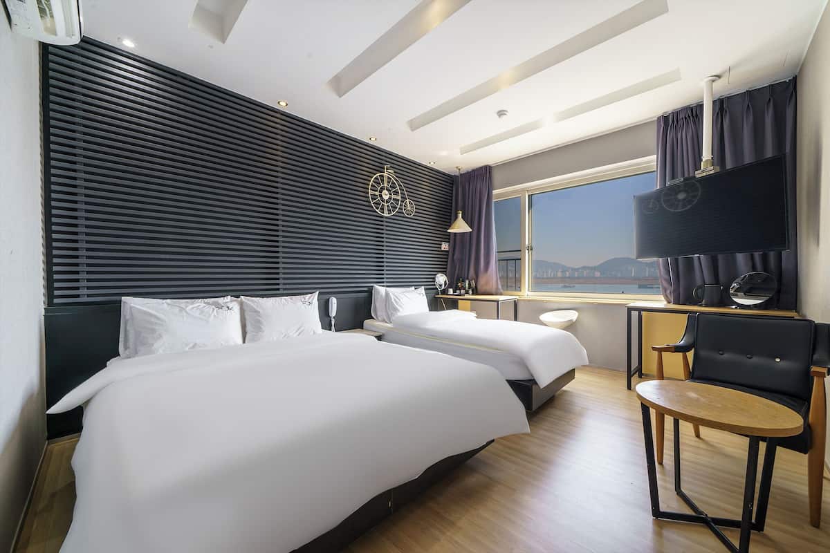 Ein Hotelzimmer mit zwei Betten, einem Schreibtisch mit Stuhl, einem Fernseher und einem Fenster mit Blick.