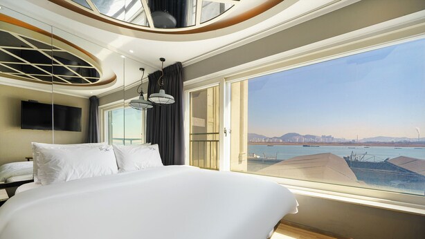 A modern hotel room with a large bed, a TV mounted on the wall, and a view of the city and water through large windows. - JK Hotel (Busan)