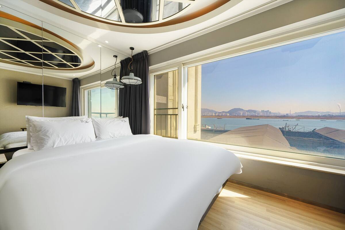 Ein modernes Hotelzimmer mit einem großen Bett, einem an der Wand montierten Fernseher und Blick auf Stadt und Wasser durch große Fenster.