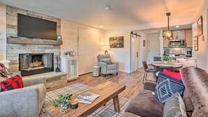 A living room with a fireplace, a flat-screen TV mounted on a stone wall, a dining area with a table and chairs, and a kitchen with modern appliances. - 'Nature Retreat' at Seventh Mountain Resort! (Bend)