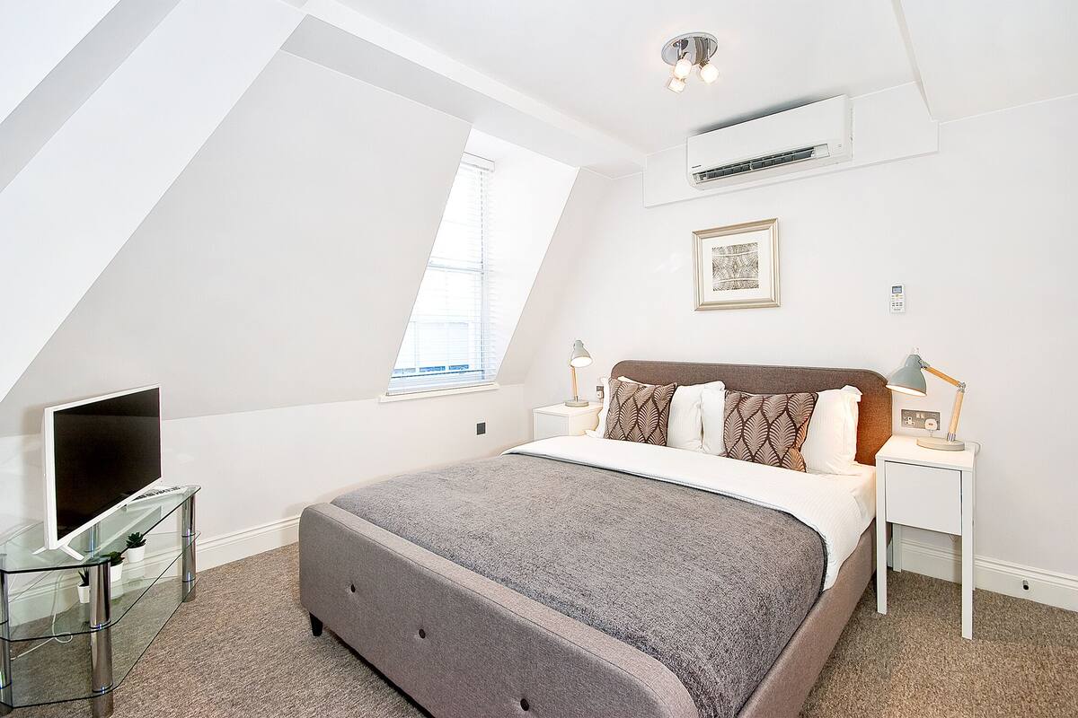 Une chambre moderne avec un grand lit, un téléviseur posé sur une table en verre et une fenêtre laissant entrer la lumière naturelle.