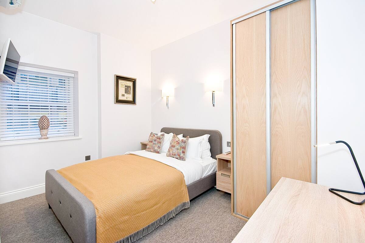 Une chambre à coucher comprenant un lit, des tables de chevet, une commode, une armoire en bois, une fenêtre avec des stores et des lampes fixées au mur.