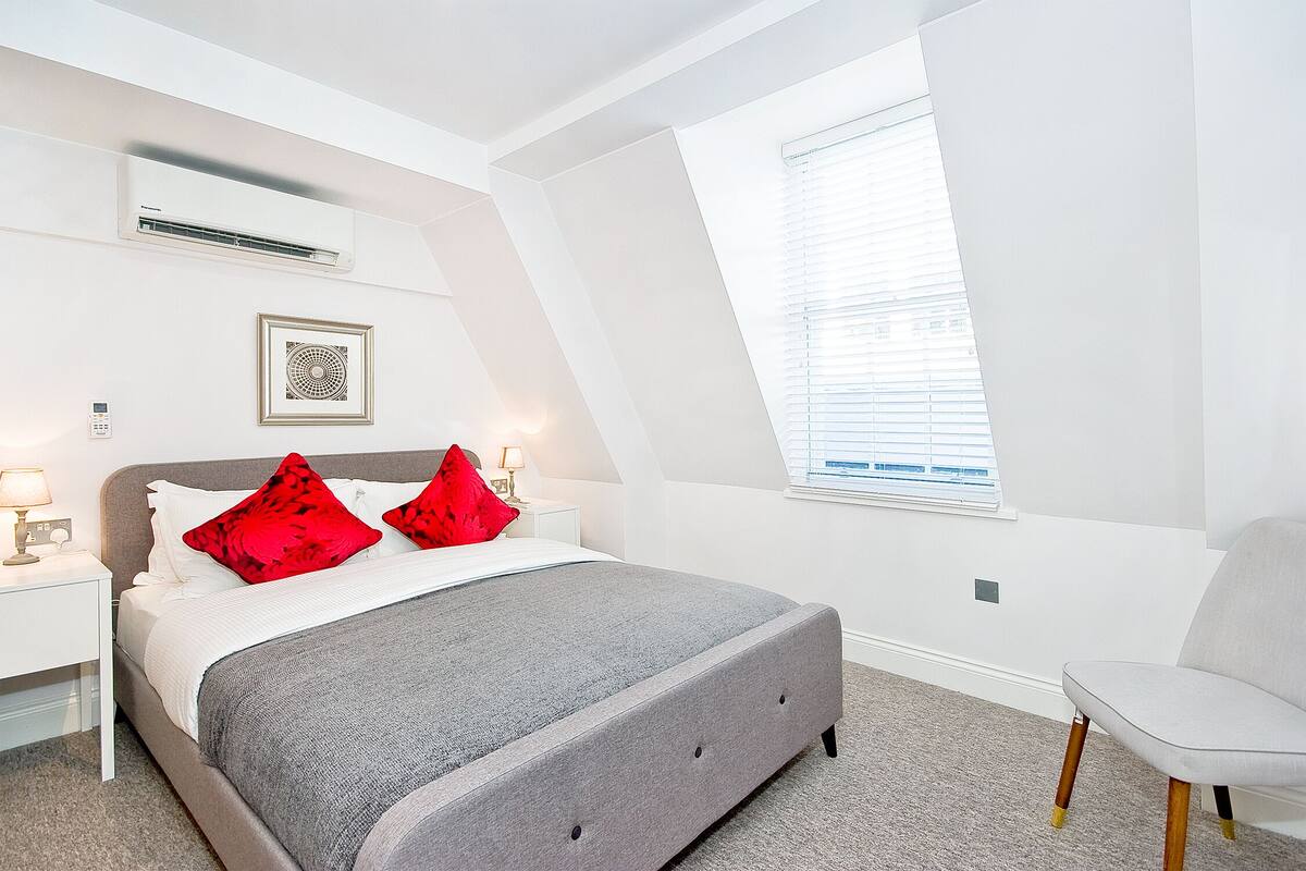 Une chambre à coucher moderne avec un lit, des oreillers rouges, une table de chevet, une lampe et une fenêtre avec des stores.