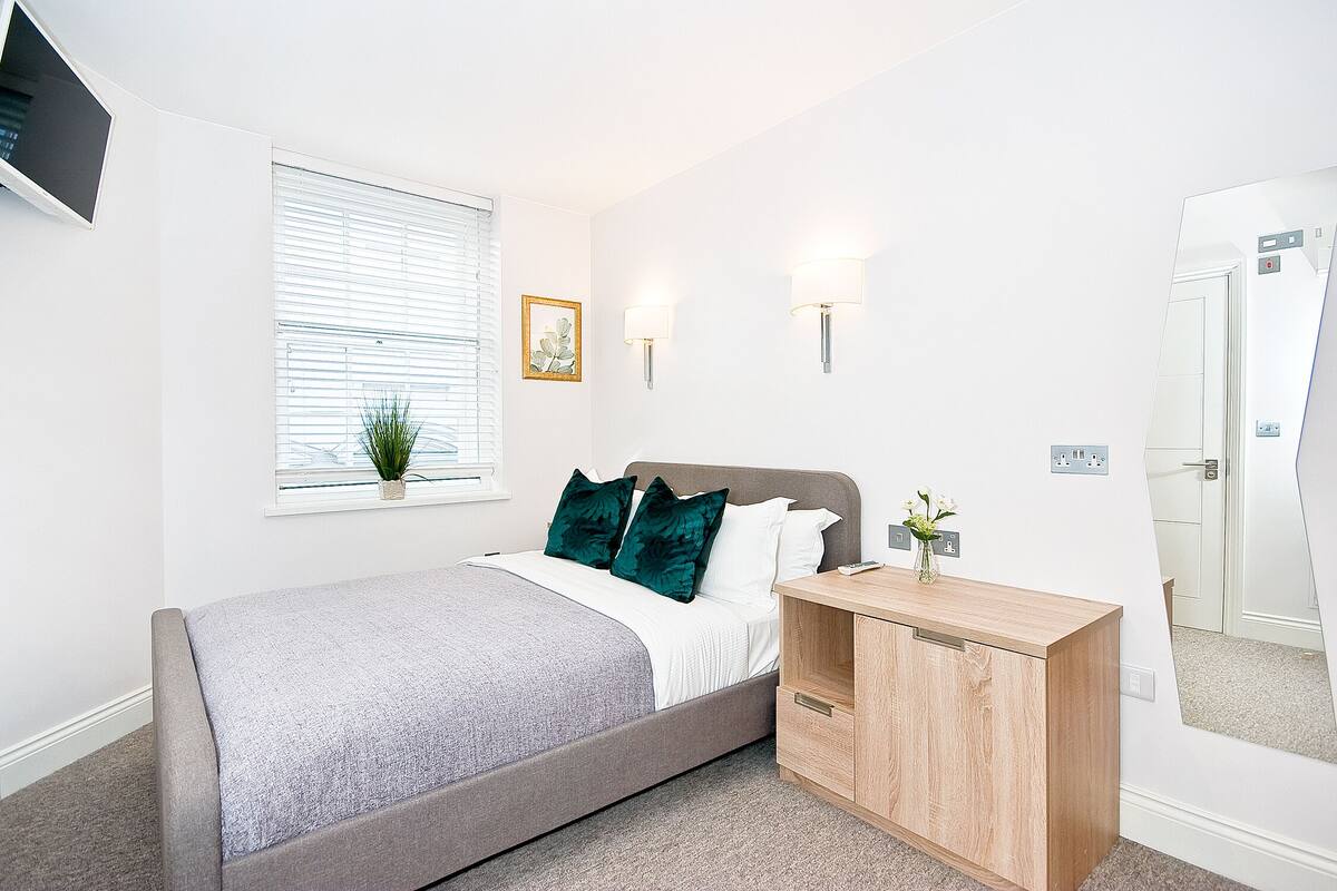 Une chambre à coucher avec un lit, une table de chevet, une lampe, une fenêtre et un miroir.