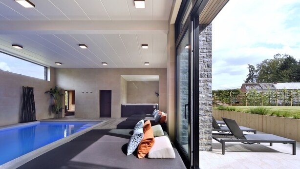 Ein moderner Poolbereich im Innenbereich mit Liegestühlen und einem großen Fenster mit Blick auf einen Garten.