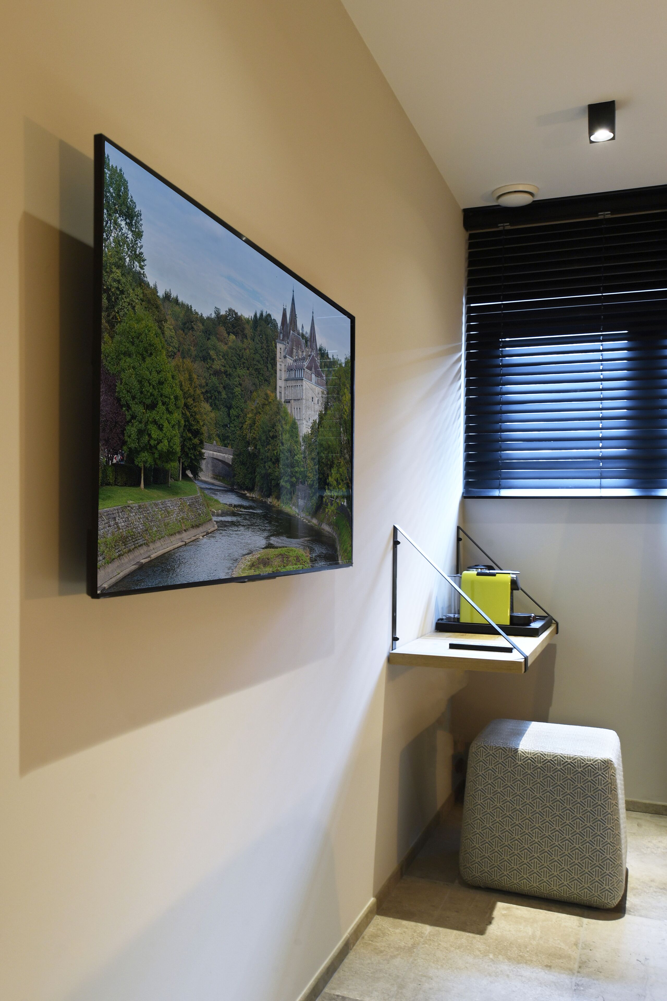 Een moderne kamer met een flatscreen televisie aan de muur, een tafeltje met een lamp en een raam met zonwering.