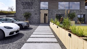 Ein modernes Gebäude mit Naturstein-Fassade und einem Schild mit der Aufschrift "Durbuy Suites". Vor dem Gebäude sind zwei Autos geparkt, und ein Kiesweg führt zum Eingang.