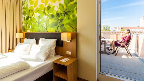 A hotel room with a bed, a nightstand, a wall-mounted lamp, and a view of a balcony with a person sitting outside. - INhouse Wohnen auf Zeit (Ingolstadt)