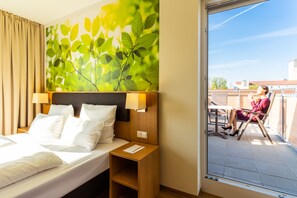Ein Hotelzimmer mit einem Bett, einem Nachttisch, einer Wandlampe und Blick auf einen Balkon mit einer sitzenden Person.