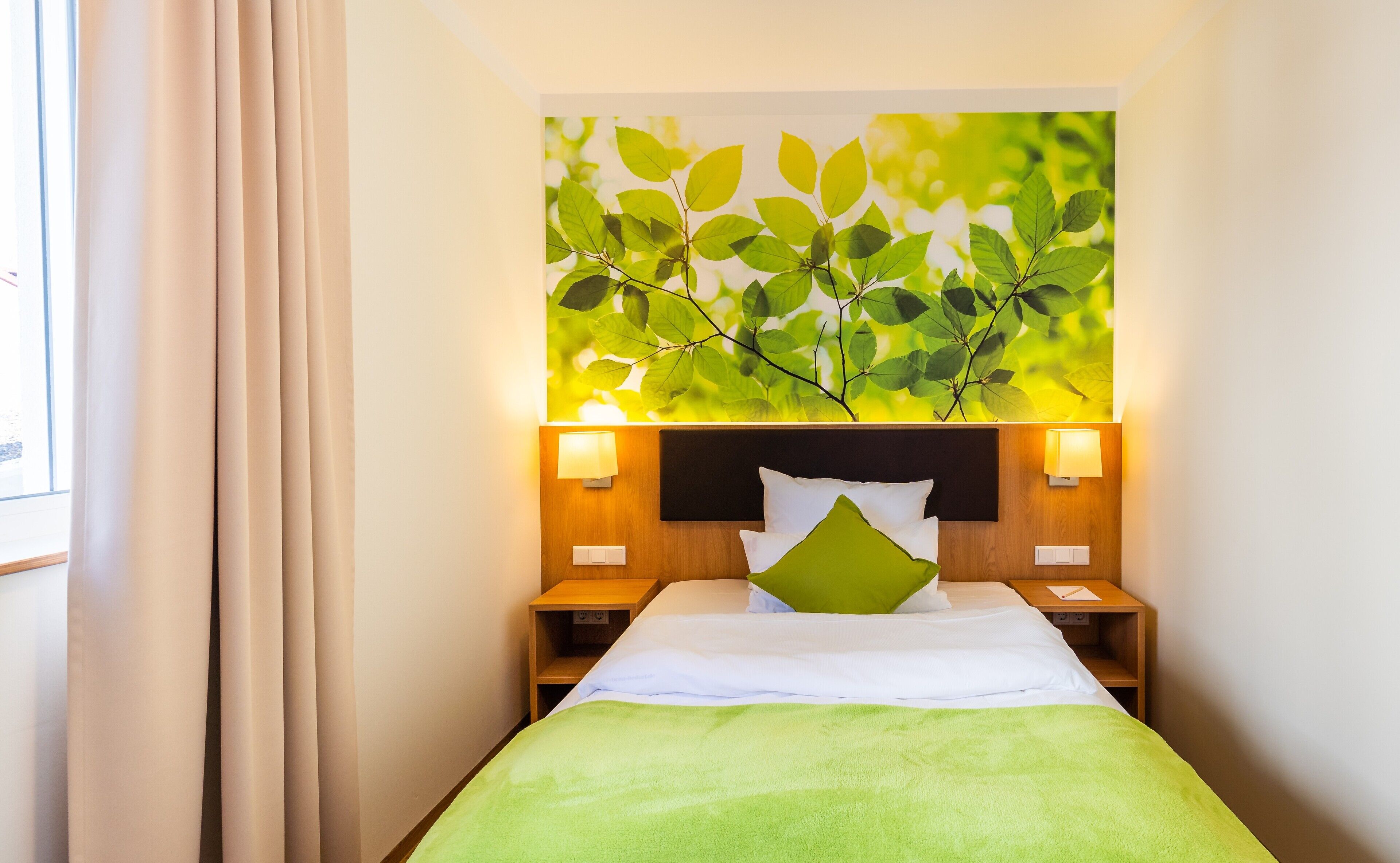 A hotel room with a bed, a green blanket, a wall-mounted artwork of leaves, and two bedside tables with lamps.