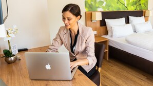 A hotel room with a bed, a desk with a laptop, a nightstand, a lamp, and a potted plant. - INhouse Wohnen auf Zeit (Ingolstadt)