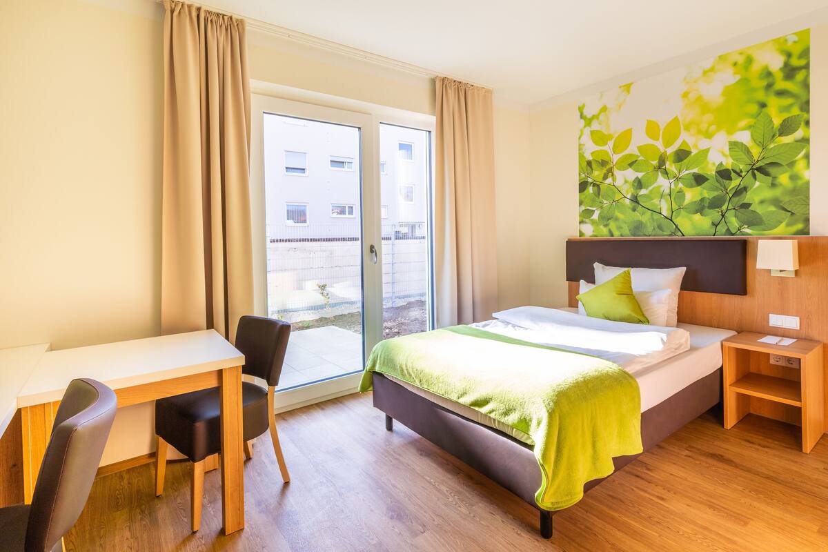 A hotel room with a bed, a desk, and a chair. There is a window with curtains, a wall with a leaf pattern, and a view of buildings outside.