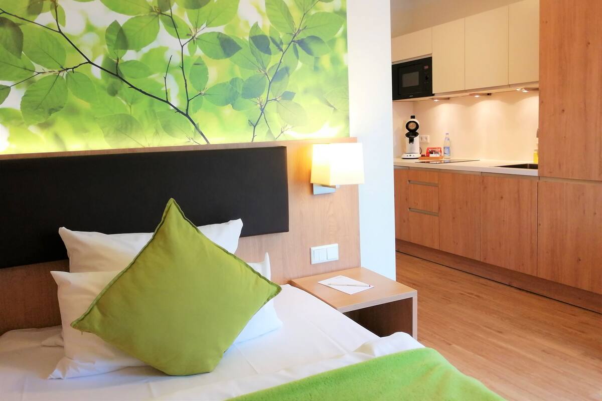 A hotel room with a bed, a green pillow, a nightstand, a wall-mounted lamp, and a view of the kitchen.