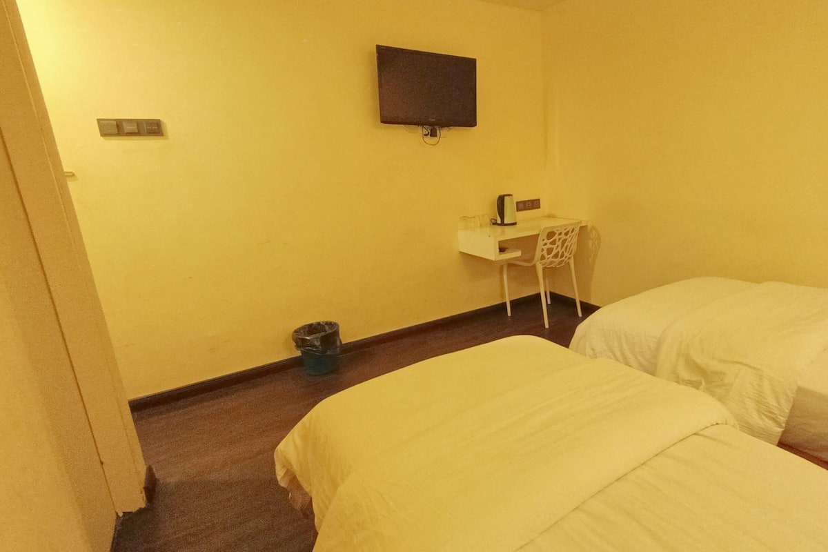 A hotel room with two beds, a small desk with a chair, a wall-mounted TV, and a trash bin.