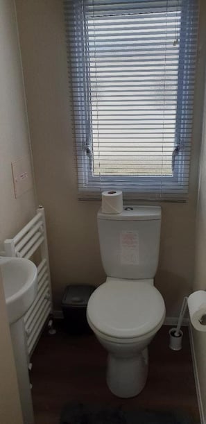 Une petite salle de bain avec des toilettes, un radiateur, une fenêtre avec des stores et un rouleau de papier toilette posé sur le radiateur.