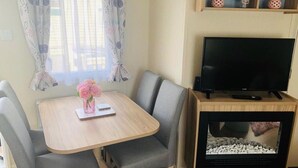 Een kamer met een televisie op een houten standaard, een open haard, een eettafel met stoelen en een raam met gordijnen.