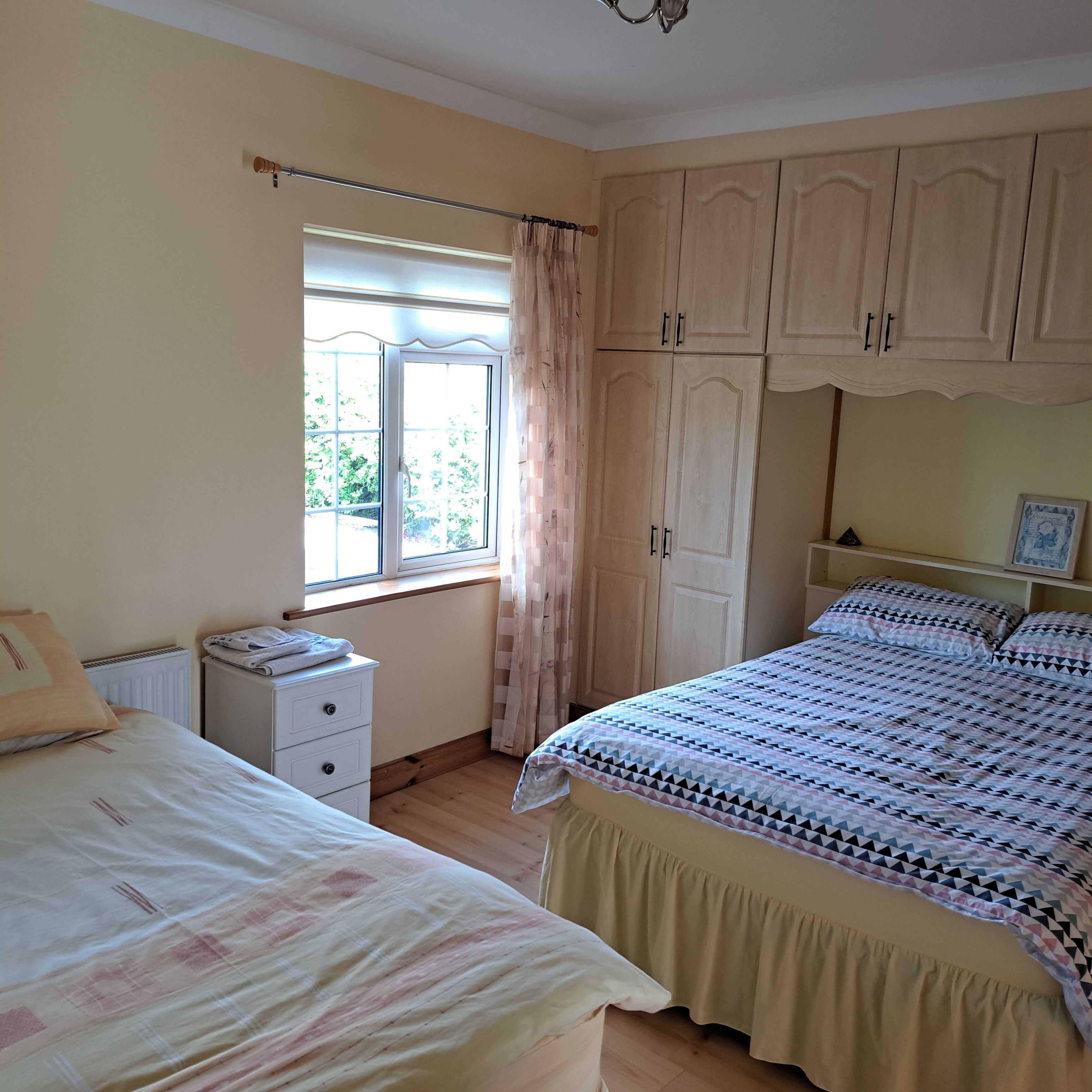 A bedroom with two beds, a window with curtains, wooden cabinets, and a radiator.