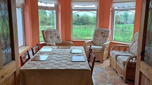 A dining area with a table set for four, surrounded by wicker chairs, large windows with a view of greenery, and a ceiling fan. - Spacious 6-bed House 10 Minutes From Knock Airport (Kilmovee)