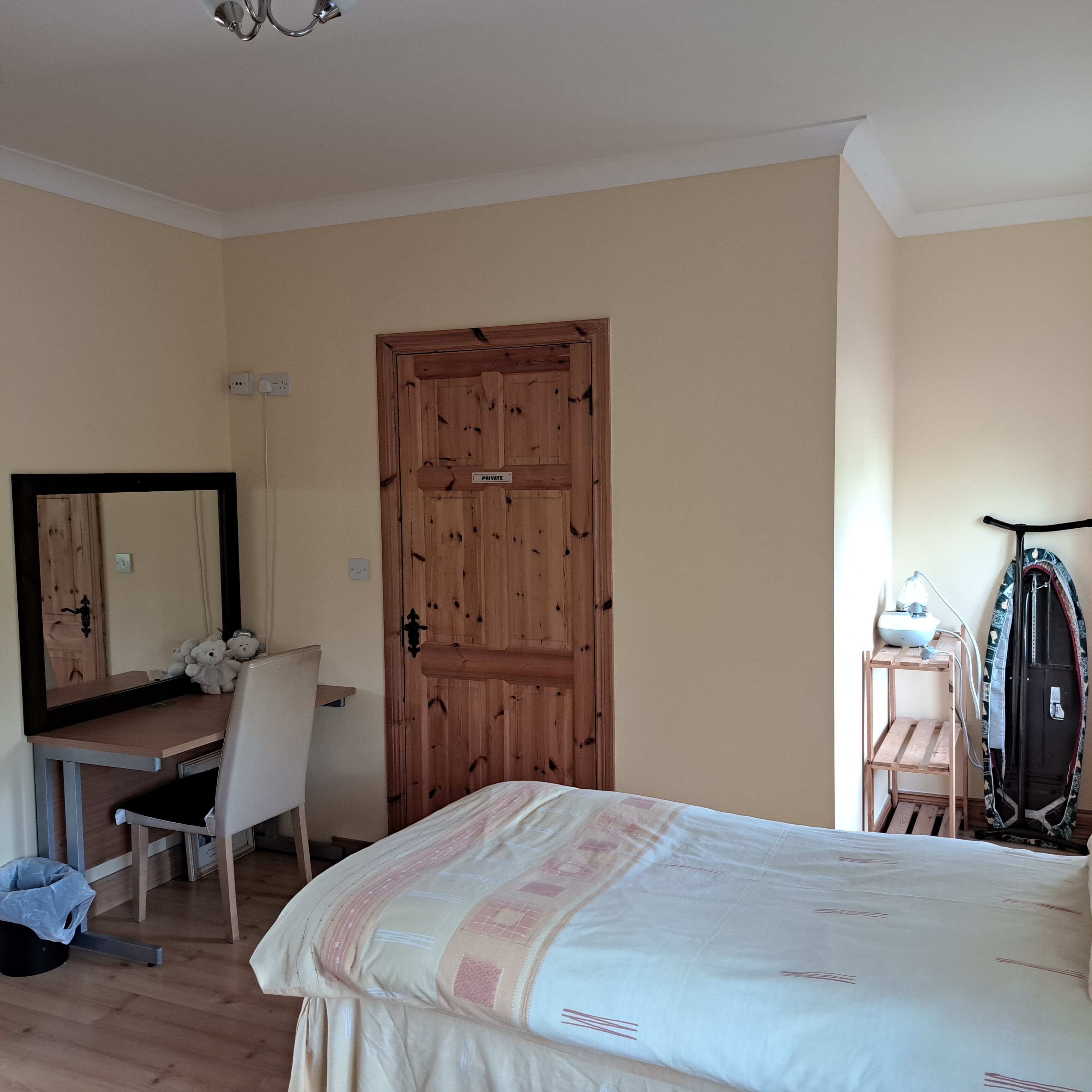 A bedroom with a wooden door, a desk with a mirror, a bed with a patterned bedspread, and a wall-mounted ironing board.