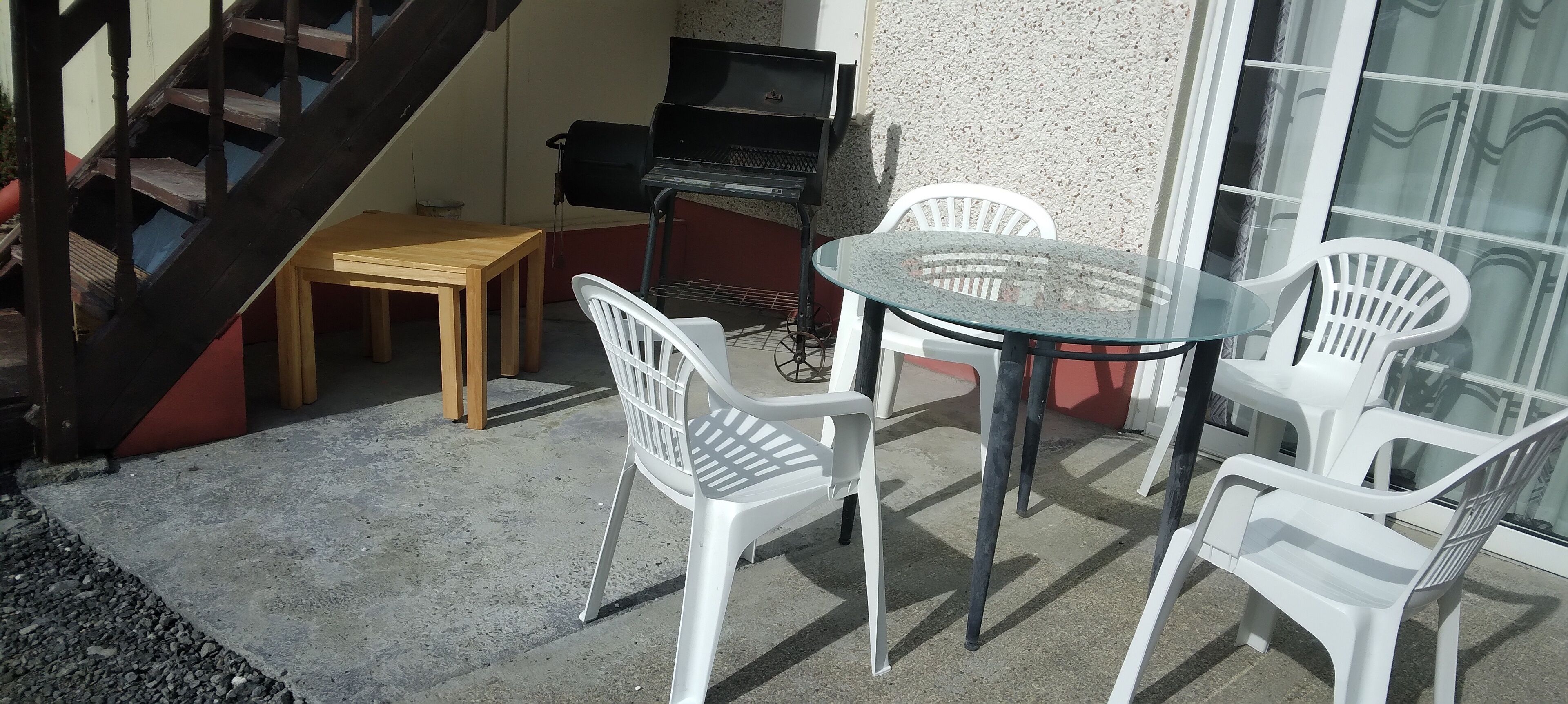 Uma área de pátio com mesa de vidro e cadeiras de plástico branco, uma mesa de madeira e uma churrasqueira.