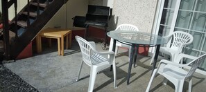 A patio area with a glass table and white plastic chairs, a wooden table, and a barbecue grill.