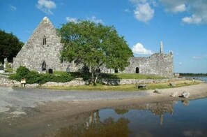 A stone building with a triangular roof, surrounded by greenery and a body of water.