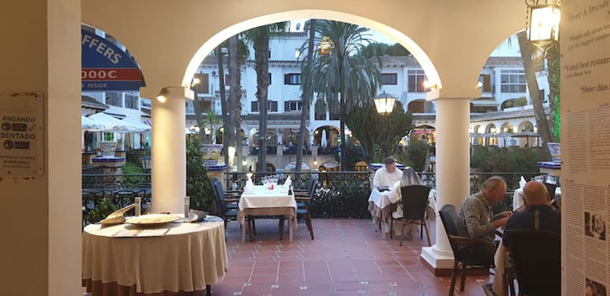 An outdoor dining area with tables and chairs, surrounded by arches and a view of buildings in the background.
