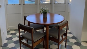 Een eetgedeelte met een ronde tafel en vier stoelen, grote ramen met uitzicht op zee en gebouwen, en een vloer met een honingraatpatroon.