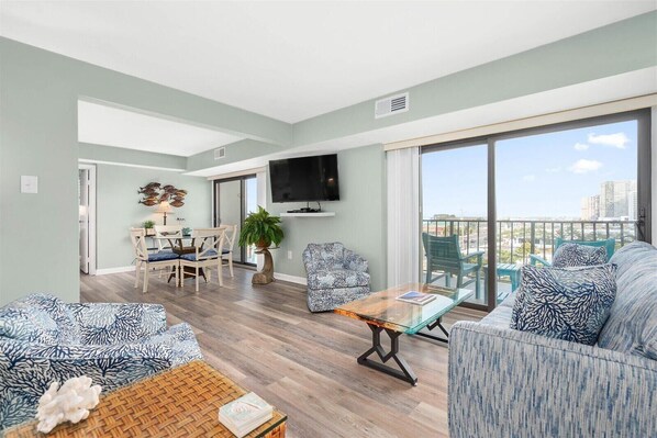 A spacious living room with a dining area, a balcony with a view, and a flat-screen TV mounted on the wall. - 9400 Building - Ocean Views in this 2 Bedroom with Outdoor Pool! (Ocean City)