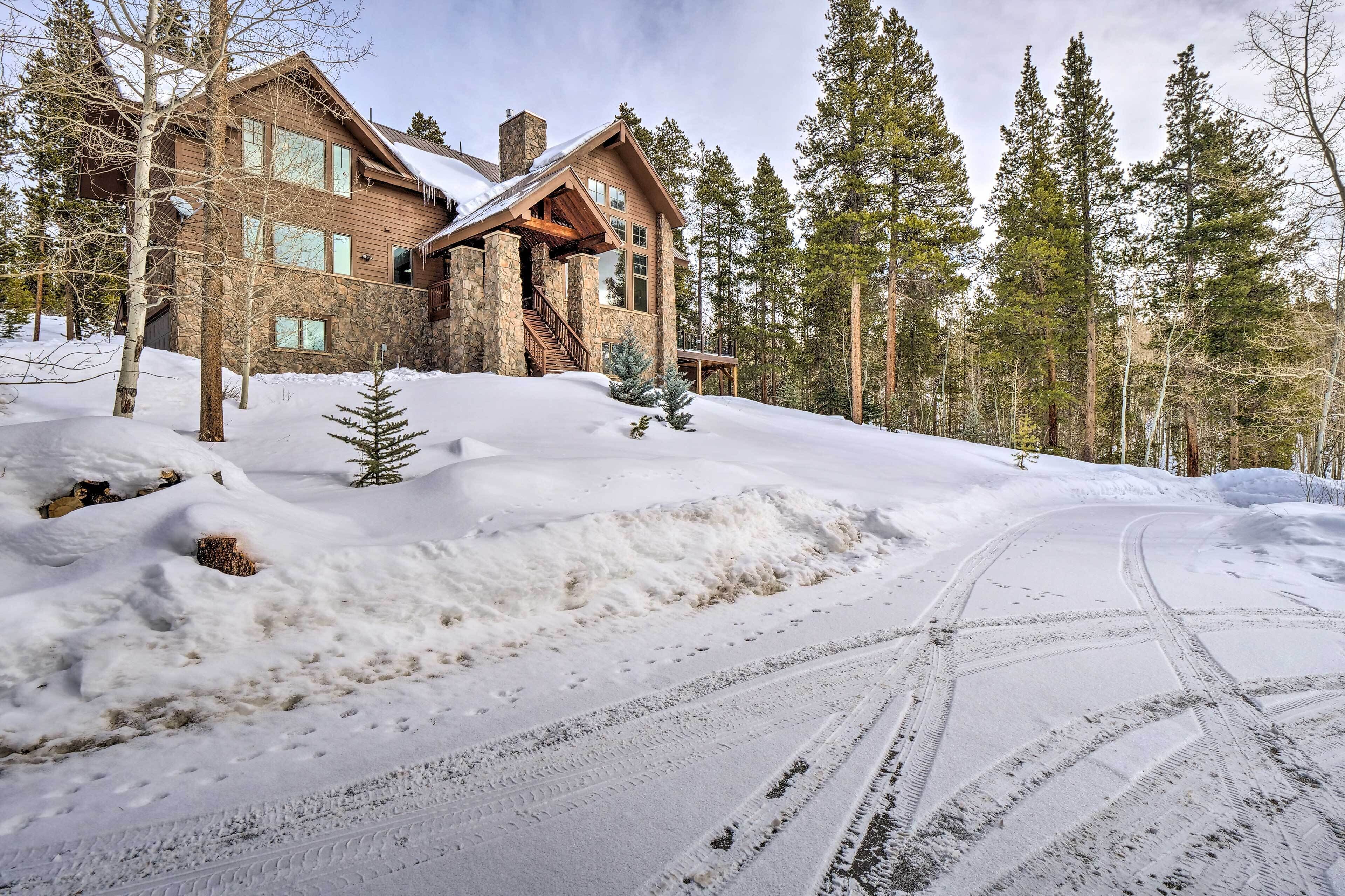A large log cabin with a stone foundation, surrounded by snow-covered ground and tall pine trees.