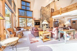 A spacious living room with large windows, a stone fireplace, and a wooden balcony.