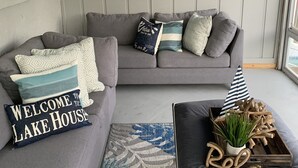 Un salon chaleureux avec un canapé d’angle gris, une petite table basse agrémentée d’une plante et un tapis. On y trouve également des coussins décoratifs et une planche de paddle fixée au mur.