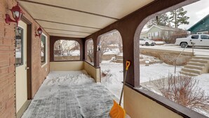 Un portico riparato con il terreno imbiancato dalla neve, una scopa appoggiata al muro e, sullo sfondo, una casa e alcuni veicoli parcheggiati.