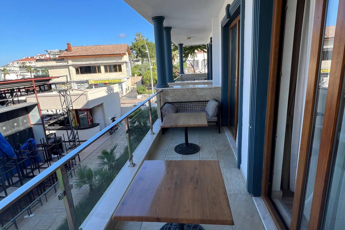 Uma varanda com um banco xadrez, uma pequena mesa e vista para a rua e edifícios.