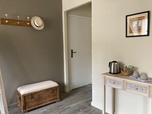 Un couloir avec un banc en bois, un porte-manteau sur lequel est posé un chapeau, une porte et une petite table avec une bouilloire et des tasses.