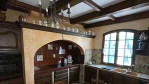 A traditional kitchen with wooden cabinets, a round table with a floral tablecloth, and a window with a view.
