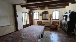 A room with a large bed, two armchairs, a wooden table, and a cabinet with decorative items.