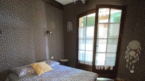 A bedroom with a patterned bedspread, a small round table, a dreamcatcher, and a window with lace curtains.