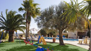 A children's play area with swings, a seesaw, and a slide, surrounded by palm trees and a pool. - Tanca - Typical Finca with Private Pool, Sleeps 8, A/C, Wi-Fi, Near Cala d'Or (Santanyí)